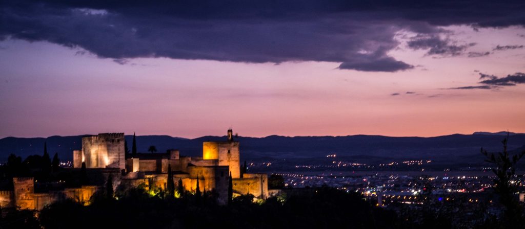 Vista impresionante de La Alhambra de Granada al atardecer, destacando sus detalladas fachadas de estilo islámico y jardines, con la ciudad de Granada y las montañas de Sierra Nevada al fondo.